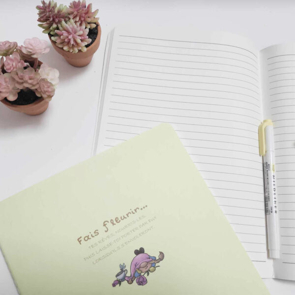 Carnet de notes cousu jaune pâle avec illustration de gnome et citation motivante, ouvert sur pages lignées avec accessoires roses